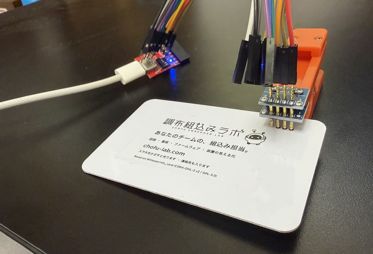 裏返した名刺基板の右上をオレンジ色のクリップではさみ、ジャンパー線で赤いアダプタにつないで、USB でパソコンに接続している